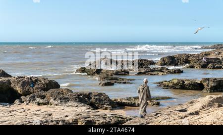 Essaouira, Marokko - 17. September 2022: Marokkanischer Mann in traditioneller Kleidung, der an der Küste von Essaouira mit Blick auf den Atlantik spaziert Stockfoto