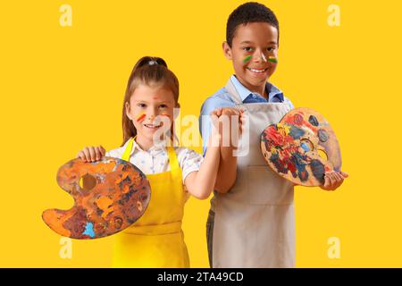 Niedliche kleine Kinder mit Gesichtern in Farben, die Hände auf gelbem Hintergrund halten Stockfoto