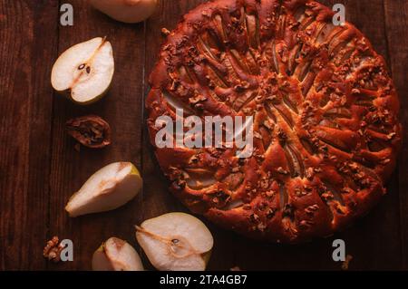 Birnenkuchen auf einem schwarzen Servierbrett auf einem Holztisch, geschnittene Birnen und Walnüsse liegen chaotisch Stockfoto