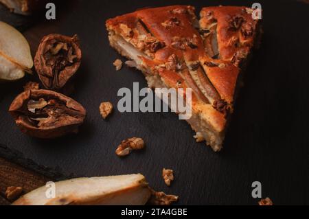 Birnenkuchen auf einem schwarzen Servierbrett auf einem Holztisch, geschnittene Birnen und Walnüsse liegen chaotisch Stockfoto