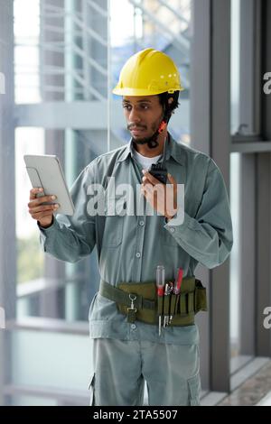 Schwarzer Vorarbeiter, der den Blueprint auf einem Tablet-Computer betrachtet und mit dem Erbauer über Walkie-Talkie kommuniziert Stockfoto