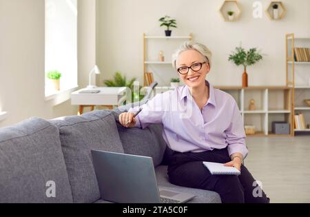 Eine schöne lächelnde Geschäftsfrau mit Brille sitzt auf dem Sofa vor dem Laptop mit Notizblock in der Hand. Stockfoto