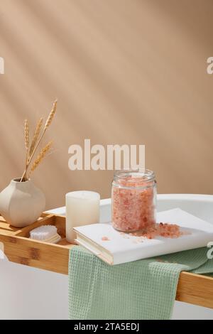 Wannentablett aus Holz mit Blumentopf, Fußbürste, Kerze, Buch und einem Glas rosa himalaya-Salz. Rosafarbenes himalaya-Salz sprengt tote Zellen weg Stockfoto