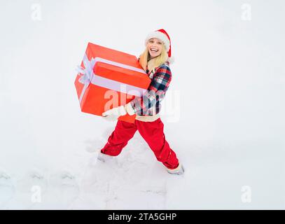 Frau trägt ein großes Geschenk isoliert auf Schnee Winter Hintergrund - voller Länge. Lustige Frau hält ein großes Weihnachtsgeschenk. Stockfoto