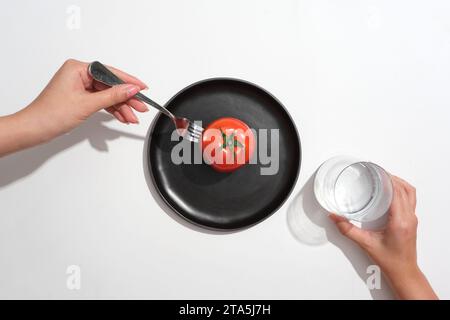 Weibliches Handmodell, das eine Gabel und ein Glas Wasser hält. Eine Tomate, die auf eine schwarze Schale gelegt wird. Das Vitamin A in Tomaten hilft, die Immunität zu stärken Stockfoto