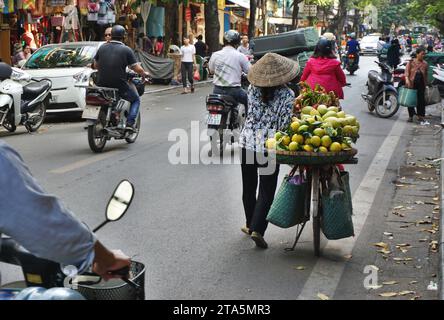Eine Obstverkäuferin mit traditionellem konischem Hut geht mit ihrem Fahrrad, beladen mit frischen Mangos und Orangen, entlang einer belebten Straße in Hanoi, Vietnam Stockfoto