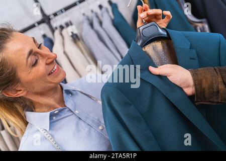 Lächelnde Segelin zeigt einem männlichen Kunden eine blaue Jacke, Kleiderständer im Hintergrund im Unkrautladen Stockfoto