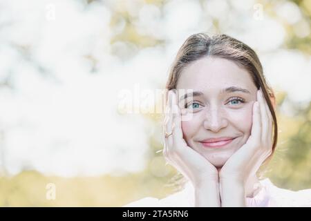 Eine junge Frau wird mit einem strahlenden Lächeln dargestellt, die Hände umrahmen ihr Gesicht und betonen ihren fröhlichen Ausdruck. Sie steht vor einem hellen und luftigen Hintergrund mit einem weichen Fokus, der das Licht und die fröhliche Atmosphäre um sie herum vermittelt. Ihre Augen funkeln vor Glück und spiegeln einen Moment der reinen Glückseligkeit und Zufriedenheit in der natürlichen Umgebung wider. Fröhlicher Blick: Strahlende Freude in der Natur. Hochwertige Fotos Stockfoto