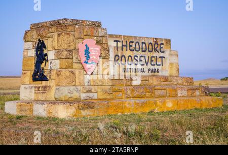 Das steinerne Eingangsschild mit der Aufschrift THEODORE ROOSEVELT NATIONAL PARK steht in trockenem Gras neben einer dunklen Metallfigur in North Dakota. Stockfoto
