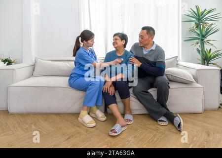 Zwei Senioren und eine Krankenschwester sitzen auf einem Sofa und nehmen an einem Gesundheitscheck in einem gemütlichen Wohnzimmer Teil. Stockfoto