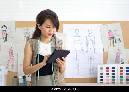 Junge asiatische Schneiderin, die E-Mail-Bestellung vom Tablet-Computer aus überprüft. Muster für Tuch, Seidenfaden Farbkatalog und verschiedene Nähartikel Stockfoto