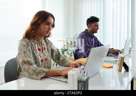 Ernsthafte junge Indianerin, die im Büro am Laptop arbeitet und E-Mails von Kunden beantwortet Stockfoto