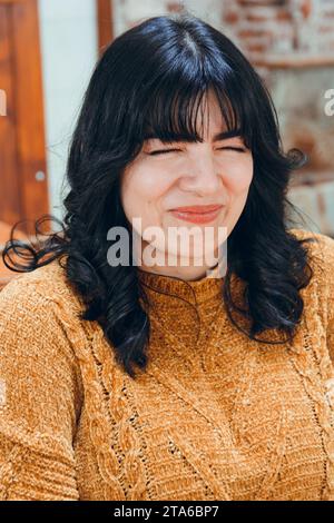 Porträt einer jungen lateinfrau mit schwarzem Haar und braunem Pullover, die mit lustigem Gesicht im Restaurant sitzt Stockfoto