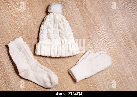 Eine weiße Strickmütze mit Pompon, Wollhandschuhen und Socken liegt auf hölzernem Hintergrund. Details zur Winterkleidung, flach geschnitten, Draufsicht Stockfoto