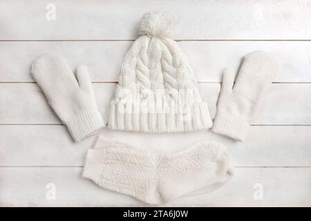 Eine weiße Strickmütze mit Pompon, Wollmittensa und Socken liegt auf weißem Holzhintergrund. Details zur Winterkleidung, flach geschnitten, Draufsicht Stockfoto