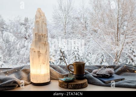 Gemütliches Winterhaus mit elektrischer Turmlampe aus natürlichem Selenit-Kristall beleuchtet auf Fensterbank, grauem Schal und Handschuhen, Papier Notizbuch, Tee. Stockfoto