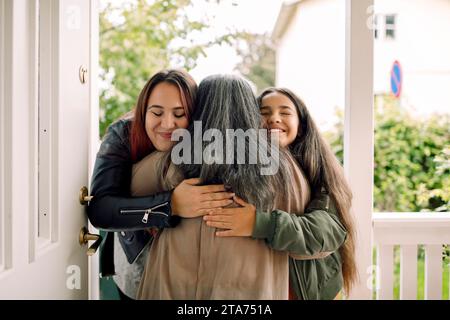 Rückansicht einer älteren Frau, die lächelnde Tochter und Enkelin am Hauseingang umschließt Stockfoto