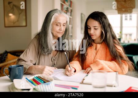 Eine ältere Frau, die Enkelin bei den Hausaufgaben hilft, während sie am Tisch sitzt Stockfoto