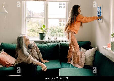 Mädchen arrangierte Gemälde an der Wand, während Großmutter zu Hause auf dem Sofa sitzt Stockfoto