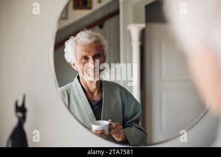 Lächelnder Seniorenmann mit Kaffeetasse, der sich zu Hause im Spiegel ansieht Stockfoto