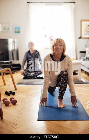 Lächelnde Seniorin, die Yoga auf einer Trainingsmatte mit einem Freund im Hintergrund zu Hause übt Stockfoto