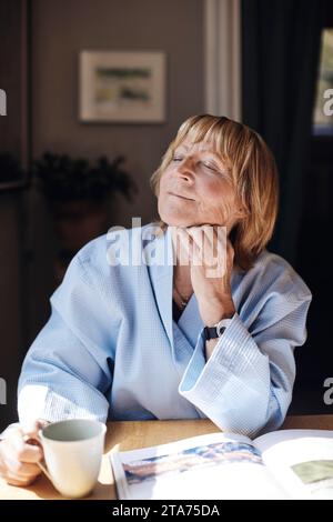 Ältere Frau mit Hand am Kinn, die eine Kaffeetasse hält, während sie sich am Tisch entspannt Stockfoto
