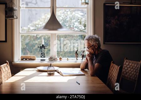 Seniorenmann mit Brille, der zu Hause ein Buch auf dem Tisch liest Stockfoto