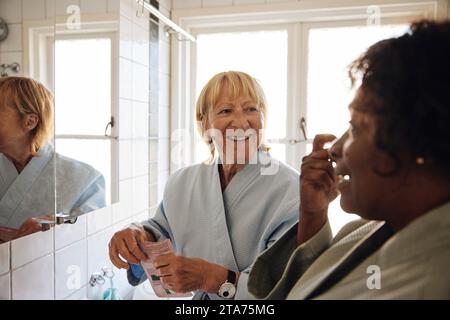 Glückliche blonde Frau, die mit einer Freundin spricht, während sie Hautpflege im Badezimmer zu Hause macht Stockfoto