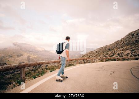 Mann mit Rucksack Skateboarden auf der Straße in der Nähe von Bergen gegen Himmel Stockfoto