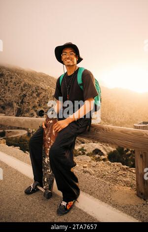 Porträt eines lächelnden jungen Mannes, der mit Skateboard auf einer hölzernen Leitplanke sitzt Stockfoto