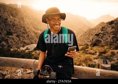 Lächelnder junger Mann, der sein Smartphone benutzt, während er sich während des Urlaubs auf das Geländer lehnt Stockfoto