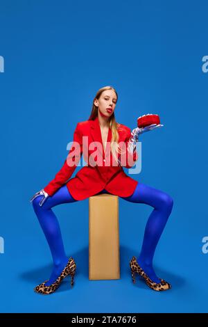 Modestandards. Junge Dame posiert in ungewöhnlichem, freakigem, hellem Outfit, trägt blaue Strumpfhosen, rote Jacke und tierbedruckte Absätze mit Geburtstagskuchen Stockfoto