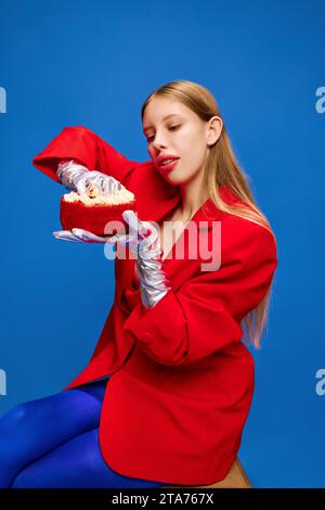 Modell der Ernährung. Ein junges Mädchen in ungewöhnlichem, freakigem, hellem Outfit, mit blauen Strumpfhosen, roter Jacke, die Geburtstagskuchen hält und mit Händen essen geht. Stockfoto