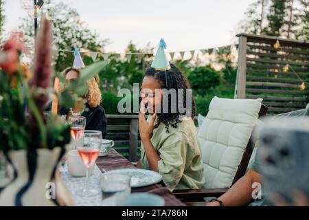 Lächelnde Frau mit Partyhut, während sie auf dem Stuhl im Garten sitzt Stockfoto
