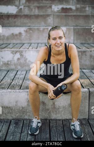 Porträt einer lächelnden Frau, die eine Wasserflasche hält, während sie auf Stufen sitzt Stockfoto