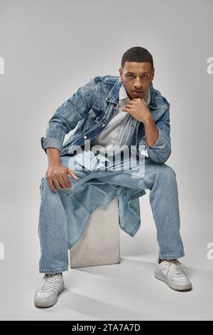 Gut aussehendes afroamerikanisches Männermodell in Denim-Outfit auf weißem Würfel, Modekonzept Stockfoto