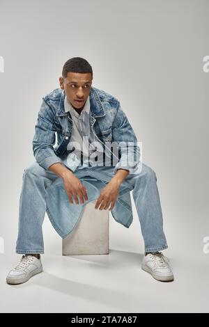 Gut aussehendes afroamerikanisches Männermodell in Denim-Outfit auf weißem Würfel, Modekonzept Stockfoto