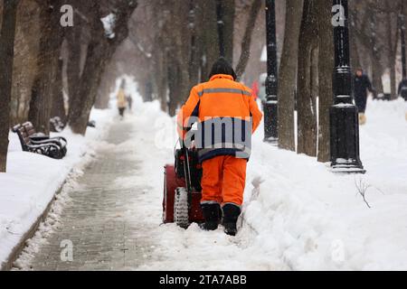 Der Gemeindedienstmitarbeiter in Uniform mit Schneepflug räumt Schnee auf einem Gehweg. Mann bei Schneeräumung in der Winterstadt, Straßenreinigung Stockfoto