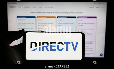 Person, die ein Mobiltelefon mit dem Logo des amerikanischen Fernsehverteilers DirecTV LLC vor der Webseite hält. Schwerpunkt auf dem Display des Telefons. Stockfoto