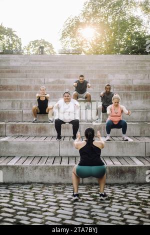 Team, das Squats auf Treppen mit weiblicher Fitnesstrainerin im Park macht Stockfoto