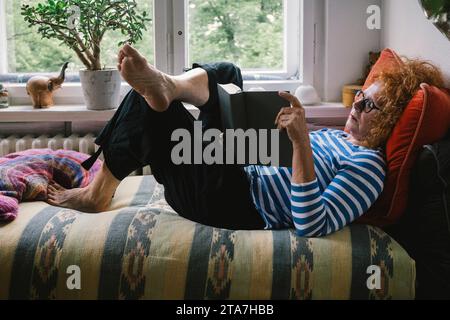 Seitenansicht einer Seniorin, die Buch liest, während sie zu Hause auf dem Bett liegt Stockfoto