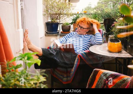 Ältere Frau liest Buch, während sie auf dem Stuhl auf dem Balkon sitzt Stockfoto