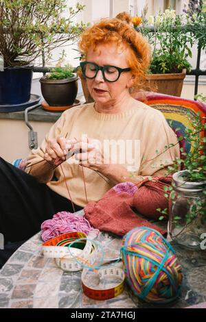 Eine ältere Frau strickt einen Pullover, während sie am Tisch auf dem Balkon sitzt Stockfoto