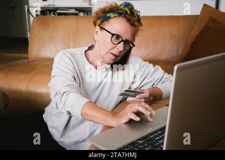 Ältere Frau, die über das Smartphone spricht, während sie Online-Zahlungen über Laptop tätigt Stockfoto