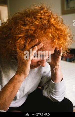 Depressive, rothaarige ältere Frau, die mit dem Kopf in den Händen auf dem Bett sitzt Stockfoto