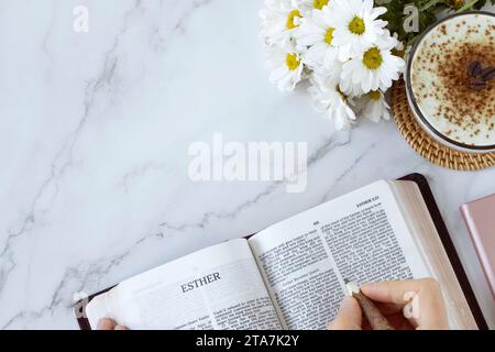 Öffnen Sie das heilige Bibelbuch von Esther mit einer Tasse Kaffee und Blumen auf weißem Marmorhintergrund. Draufsicht, Kopierraum. Das Studium Der Alttestamentlichen Schriften. Stockfoto