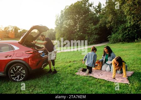 Die Familie bereitet sich auf ein Picknick vor, während sie auf dem Gras in der Nähe des Parkplatzes sitzt Stockfoto