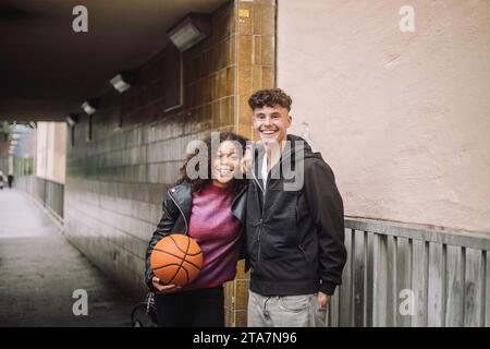 Porträt eines lächelnden Mädchens, das Basketball hält, während er mit einem männlichen Freund in der Nähe der Unterführung steht Stockfoto