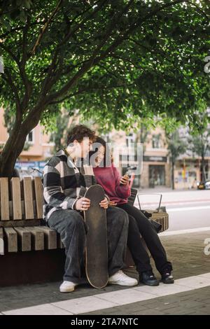 Ein lächelndes Mädchen teilt sich das Smartphone mit einem männlichen Teenager-Freund, der auf einer Bank unter dem Baum sitzt Stockfoto