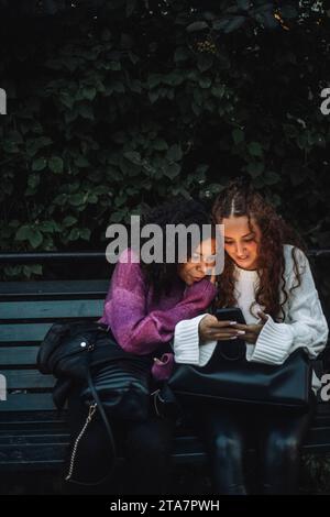 Neugierige Freundinnen, die ihr Smartphone teilen, während sie auf einer Bank in der Nähe von Pflanzen sitzen Stockfoto